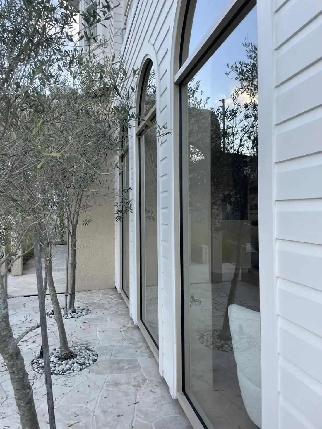 Tall arched windows on a white weatherboard home with olive trees out front